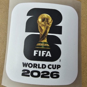 Può includere: Un adesivo quadrato con angoli arrotondati che presenta il testo nero 26, con il trofeo dorato della Coppa del Mondo FIFA che sostituisce lo zero. Sotto, il testo recita FIFA WORLD CUP 2026 in lettere maiuscole nere su sfondo bianco.