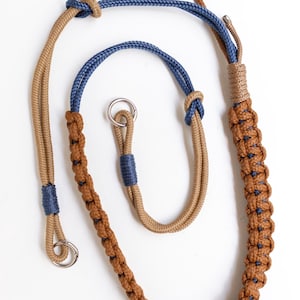May include: A braided rope leash featuring tan and blue cordage with intricate knotting patterns. The design includes two silver-toned metal rings at the ends, secured with tight blue and tan thread wraps, creating a durable and functional accessory.