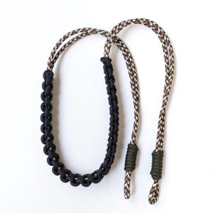 May include: A lanyard crafted from paracord featuring a central section of black braided cord. The upper portion consists of a brown, black, and white camouflage patterned cord, with two dark olive green knots securing the ends of the braid.