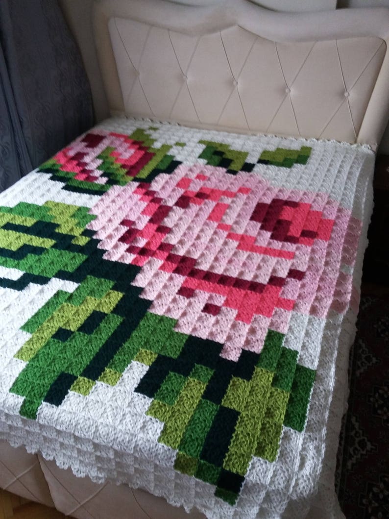 May include: A white crocheted blanket with a large pink and red rose design. The rose is surrounded by green leaves.