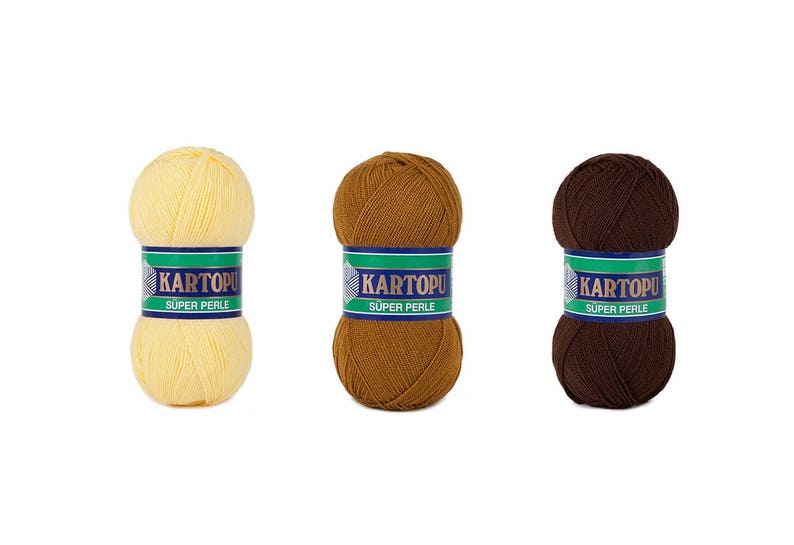 May include: Three balls of KARTOPU Super Perle yarn in shades of yellow, brown, and dark brown. Each ball is wrapped with a label featuring the brand name and product details. The yarn is likely used for knitting or crochet projects.