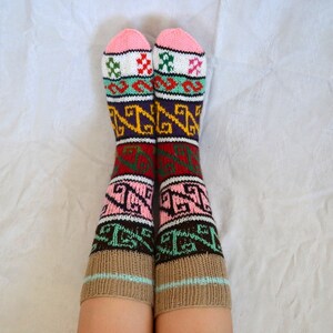 May include: A pair of colorful knitted socks with a geometric pattern in shades of pink, brown, green, white, and purple. The socks are worn on a person's legs.