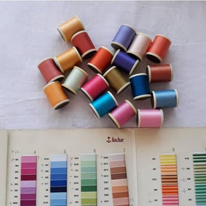 Könnte beinhalten: Eine Ansammlung bunter Garnrollen in Orange, Rot, Pink, Blau und Lila, angeordnet auf einer weißen Oberfläche. Eine Farbkarte mit dem Anchor-Logo ist sichtbar und dient als Referenz für die Garnfarben. Ideal für Näh- und Bastelprojekte.