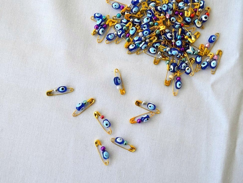 May include: A pile of gold safety pins with blue and white evil eye beads. The pins are scattered on a white fabric surface.