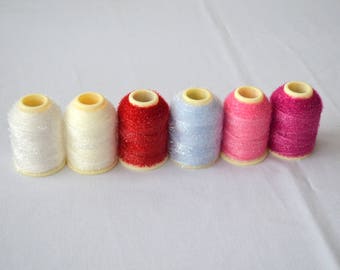 May include: Seven spools of thread in various colours, including white, red, pink, and blue. The spools are arranged in a row on a white surface.