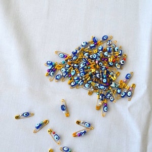 May include: A large group of gold safety pins with blue and white evil eye beads. There are also a few safety pins with colourful beads. The safety pins are scattered on a white fabric surface.