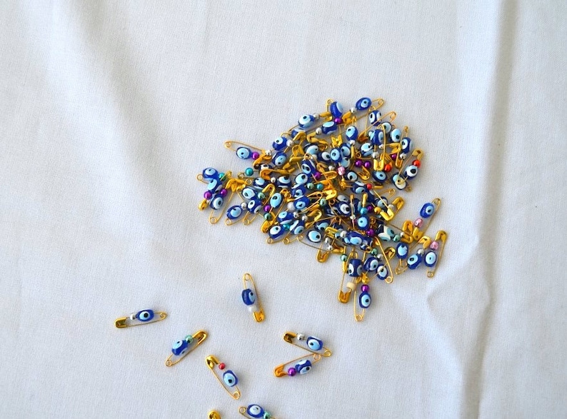 May include: A large group of gold safety pins with blue and white evil eye beads. There are also a few safety pins with colourful beads. The safety pins are scattered on a white fabric surface.