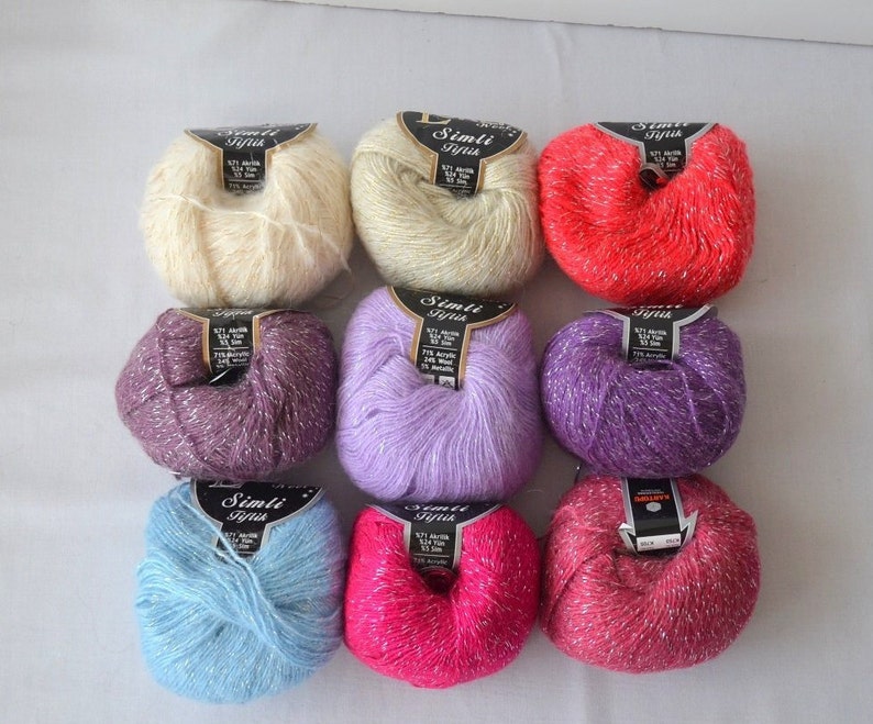 Puede incluir: Nueve ovillos de hilo con hilo de purpurina plateado. El hilo es de color crema, morado, lavanda, azul claro, rosa y rojo. Las etiquetas del hilo dicen "Simli Iplik" en texto negro.