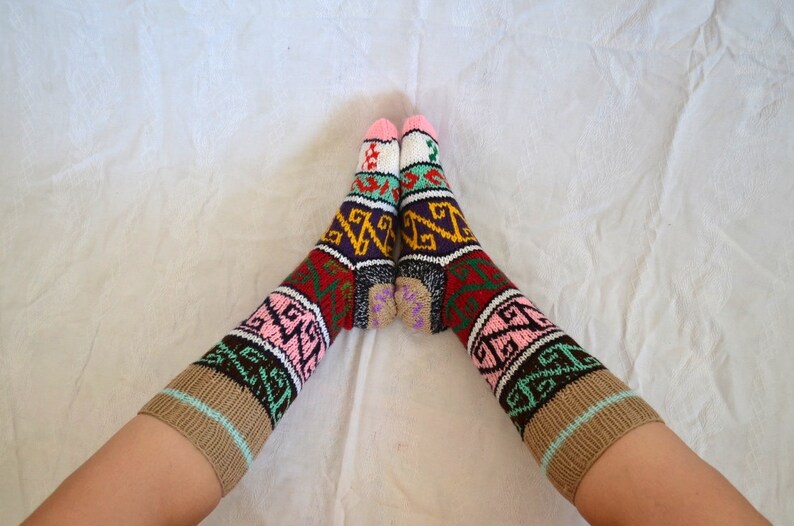 May include: A pair of colorful knitted socks with a geometric pattern in shades of brown, pink, green, red, white, and purple. The socks are folded over at the top and are worn on a person's feet.