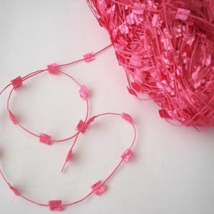 May include: Pink yarn with square-shaped embellishments. The yarn is wound into a ball and a length of the yarn is laid out straight.