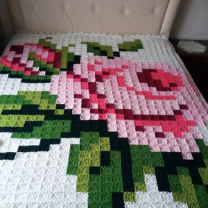 May include: A white crocheted blanket with a large pink and green rose design. The rose is made up of individual squares in different shades of pink and green.