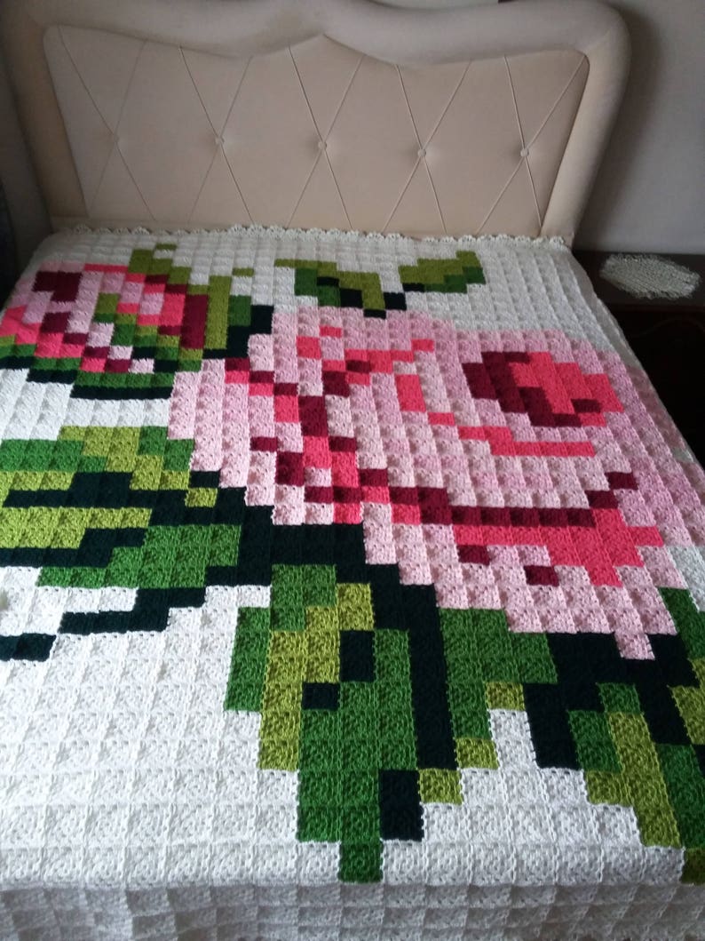 May include: A white crocheted blanket with a large pink and green rose design. The rose is made up of individual squares in different shades of pink and green.
