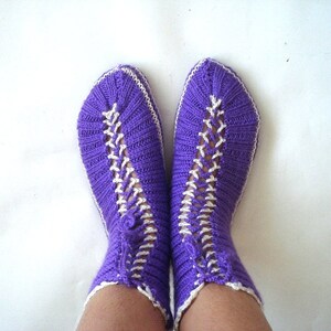 May include: Purple knitted slippers with white trim. The slippers have a criss-cross pattern on the front.