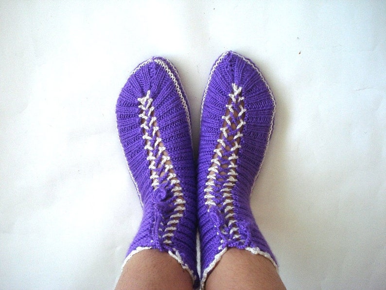 May include: Purple knitted slippers with white trim. The slippers have a criss-cross pattern on the front.