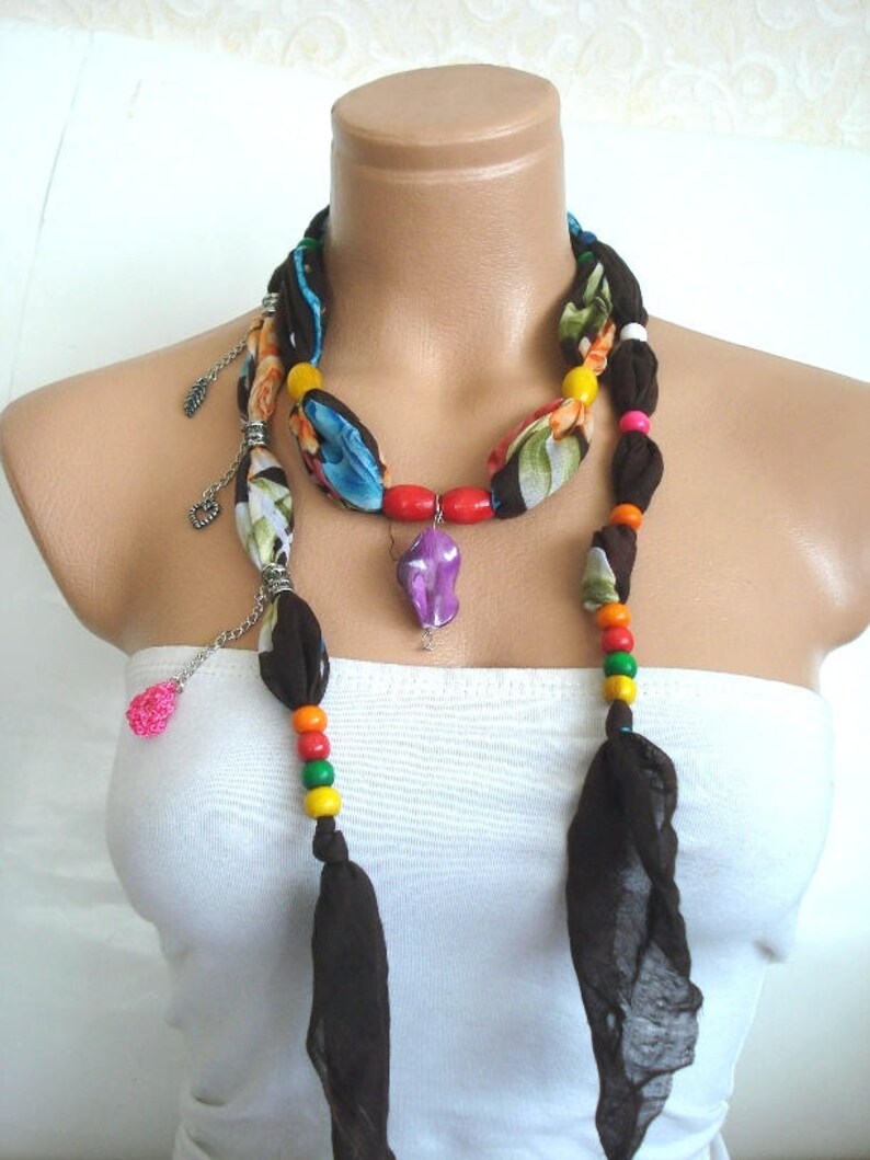 May include: A brown fabric necklace with colorful beads and a purple pendant. The necklace is layered with a floral patterned fabric and silver chain accents.