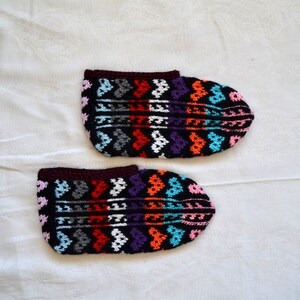 May include: A pair of colorful crocheted slippers with a geometric pattern. The slippers are black with red, orange, blue, pink, white, and purple accents.