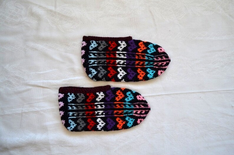 May include: A pair of colorful crocheted slippers with a geometric pattern. The slippers are black with red, orange, blue, pink, white, and purple accents.