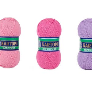 May include: Three balls of yarn in shades of pink and purple. Each ball has a label with the text "KARTOPU SUPER PERLE" in white on a green and blue background. The yarn is tightly wound, showing the texture of the fibers.