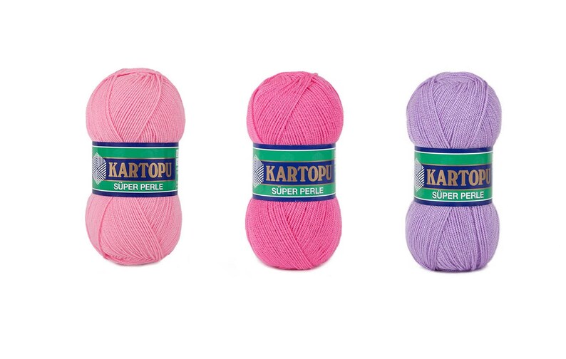 May include: Three balls of yarn in shades of pink and purple. Each ball has a label with the text "KARTOPU SUPER PERLE" in white on a green and blue background. The yarn is tightly wound, showing the texture of the fibers.