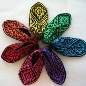 May include: A collection of seven colorful, knitted slippers arranged in a circular pattern. Each slipper features a geometric design in a different color, including orange, red, yellow, green, blue, purple, and pink, with black accents.