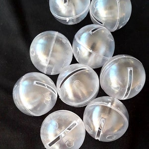 May include: A group of ten clear plastic jingle bells with a slit cut into each bell. The bells are arranged in a cluster on a black surface.