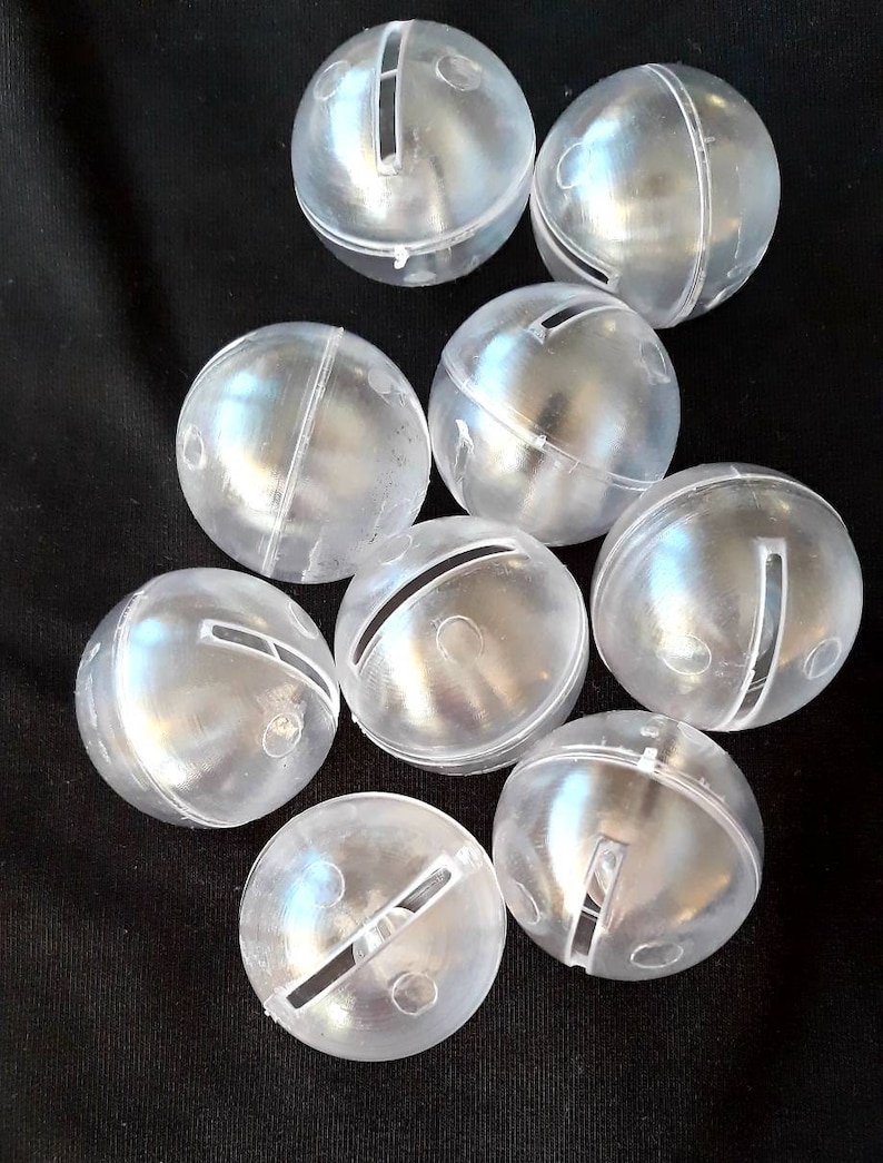 May include: A group of ten clear plastic jingle bells with a slit cut into each bell. The bells are arranged in a cluster on a black surface.