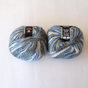 Puede incluir: Dos ovillos de hilo en un esquema de colores azul, blanco y verde con hilos met&aacute;licos plateados. El hilo est&aacute; etiquetado como "Gifiak" y "571 Acr&iacute;lico 524 Hilo 50 gr 125 m 75% Acr&iacute;lico 25% Metal."