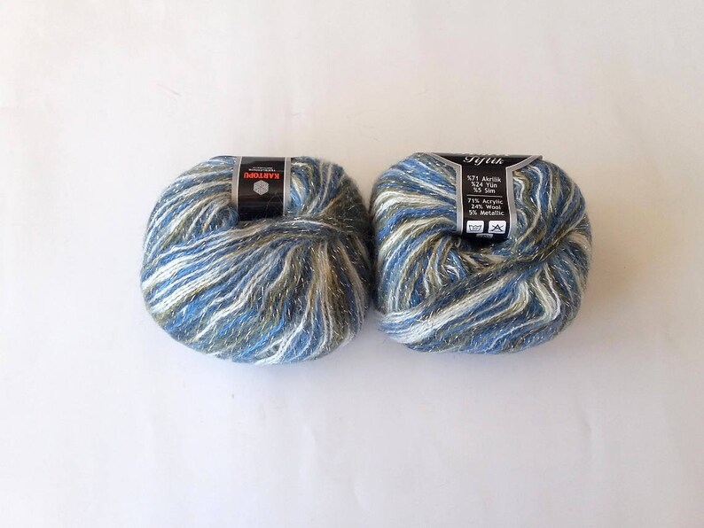 Puede incluir: Dos ovillos de hilo en un esquema de colores azul, blanco y verde con hilos met&aacute;licos plateados. El hilo est&aacute; etiquetado como "Gifiak" y "571 Acr&iacute;lico 524 Hilo 50 gr 125 m 75% Acr&iacute;lico 25% Metal."