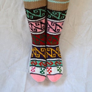 May include: A pair of colorful knitted socks with a geometric pattern. The socks are pink, white, green, brown, purple, and yellow. The socks are folded down at the top, revealing a brown cuff with a light blue stripe.
