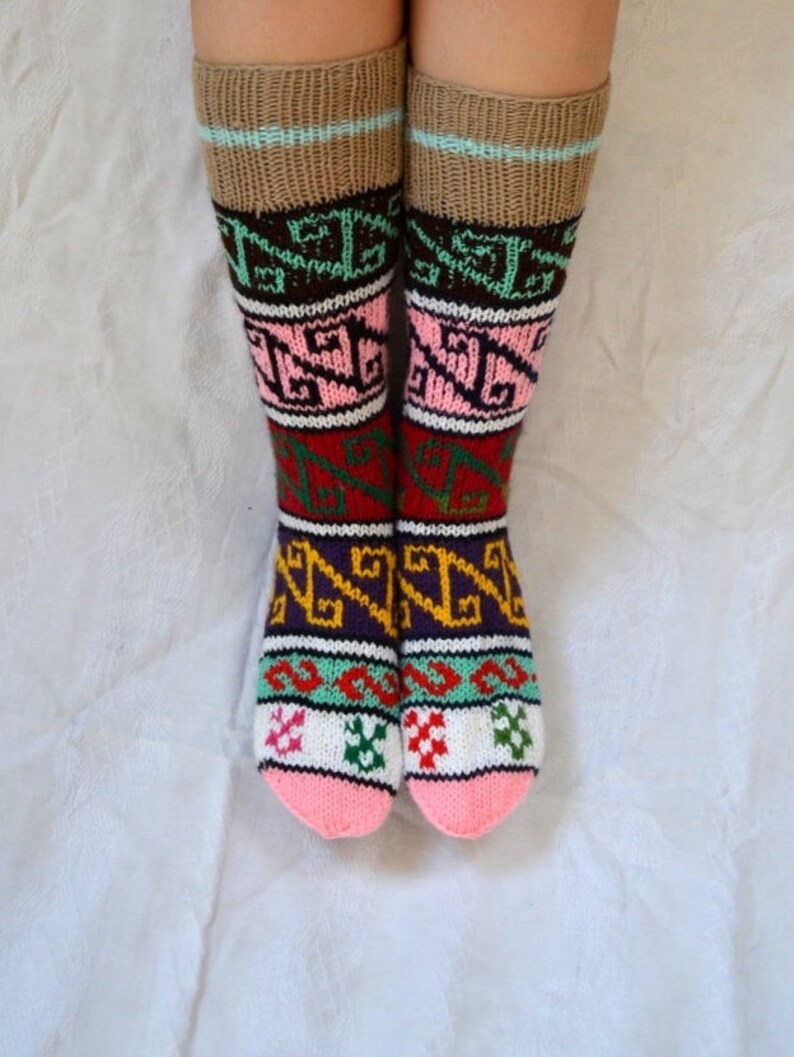 May include: A pair of colorful knitted socks with a geometric pattern. The socks are pink, white, green, brown, purple, and yellow. The socks are folded down at the top, revealing a brown cuff with a light blue stripe.