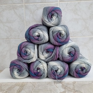 May include: A stack of ten balls of yarn in a gradient of grey, white, and purple. The yarn is arranged in a pyramid shape.