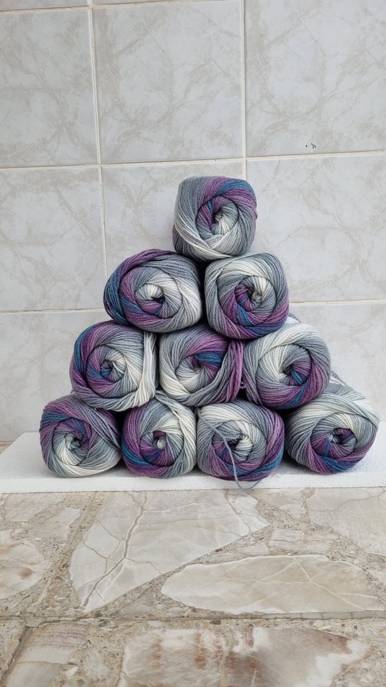 May include: A stack of ten balls of yarn in a gradient of grey, white, and purple. The yarn is arranged in a pyramid shape.