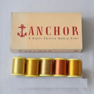 Pode incluir: Uma caixa de fio de bordar Anchor 3 Katli Ekstra Nakis Simi. A caixa é marrom com letras vermelhas e um logotipo de âncora. A caixa contém seis carretéis de fio de bordar dourado, cobre e iridescente.