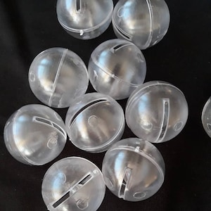 May include: A group of clear plastic jingle bells with a silver colored line across the middle. The bells are scattered on a black surface.