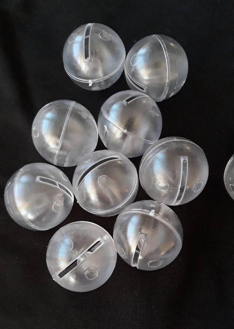 May include: A group of clear plastic jingle bells with a silver colored line across the middle. The bells are scattered on a black surface.