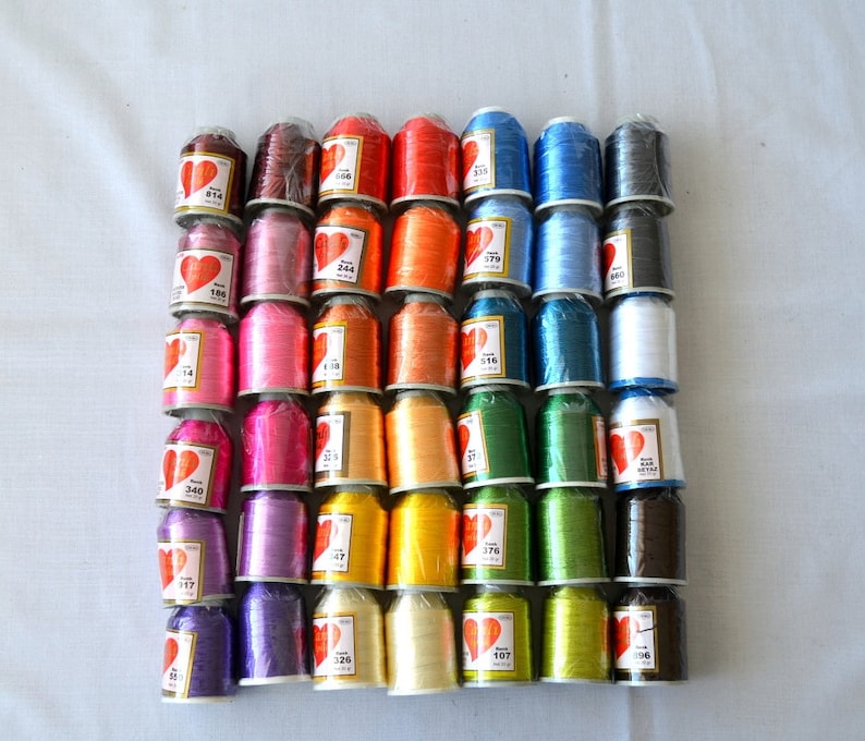 May include: A collection of 36 spools of thread in various colors, including red, orange, yellow, green, blue, purple, pink, white, and black. Each spool has a heart-shaped label with a number printed on it.