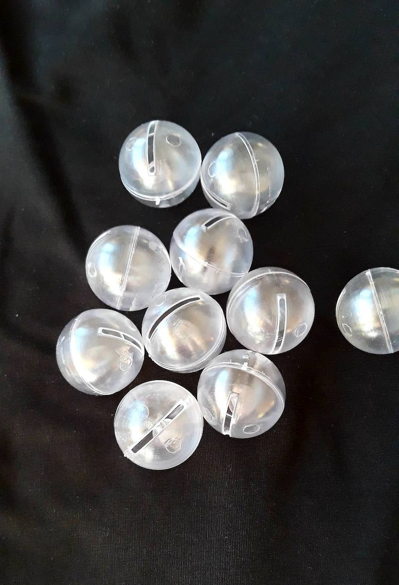May include: A group of ten clear plastic jingle bells with a single slit in each. The bells are scattered on a black surface.