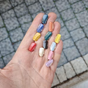 May include: A collection of 12 small, colorful, oval-shaped glass buttons with a wavy pattern. The buttons are arranged in a row on a hand.