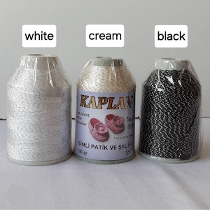 May include: Three spools of yarn in white, cream, and black. Each spool is labeled "KAPLAN" and has the text "SIMLİ PATİK VE ŞAL İPİ 100 gr 301" on the label.