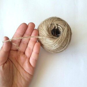 May include: A ball of natural twine, made from jute, is shown on a white surface. A hand is holding a length of twine that is unwinding from the ball.