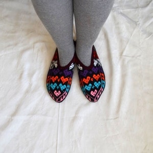 May include: A pair of colorful knitted slippers with a heart pattern in shades of purple, orange, blue, pink, and white. The slippers are worn on a white surface.