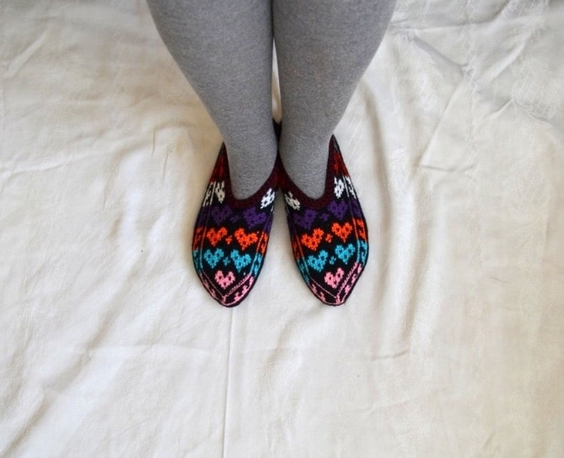 May include: A pair of colorful knitted slippers with a heart pattern in shades of purple, orange, blue, pink, and white. The slippers are worn on a white surface.
