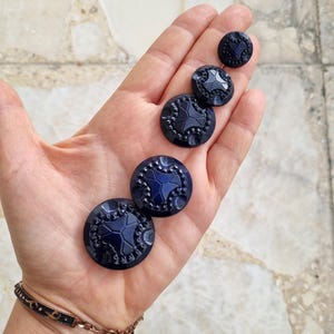 Op de afbeelding: Een verzameling marineblauwe knopen in verschillende maten. De ronde knopen hebben een stervormig patroon in het midden en een decoratieve rand. Gemaakt van glanzend materiaal.
