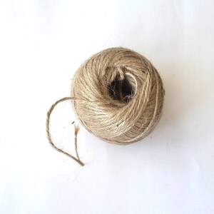 May include: A ball of natural twine, made from jute, is shown on a white background. The twine is brown and has a rough texture. The twine is unraveled and the end is visible.