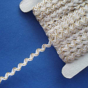 May include: White zig zag trim with a gold metallic thread running through it. The trim is wound around a white cardboard spool.