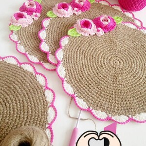 May include: Three crocheted placemats with a pink and white border and pink flowers. The placemats are made of a natural fiber, possibly jute or burlap. There is a ball of yarn and a crochet hook in the image.