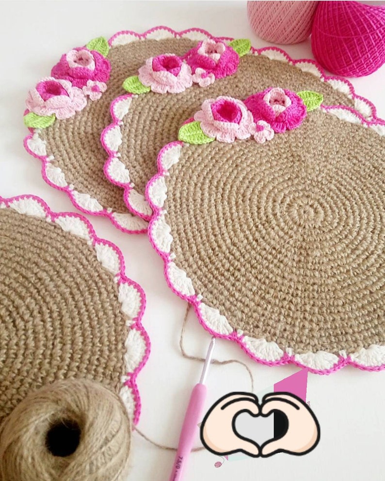 May include: Three crocheted placemats with a pink and white border and pink flowers. The placemats are made of a natural fiber, possibly jute or burlap. There is a ball of yarn and a crochet hook in the image.