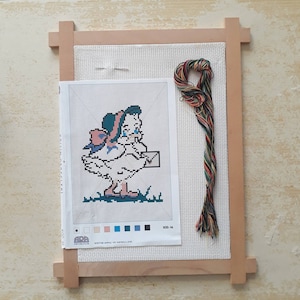 May include: A cross-stitch pattern featuring a cartoon chick wearing a pink bow and a blue hat, holding a letter. The pattern is in a wooden frame with a skein of colorful embroidery floss.