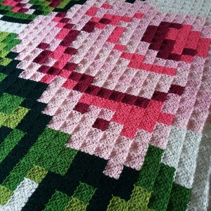 May include: A crocheted blanket with a pixelated design of a pink flower with green leaves. The blanket is made with different shades of pink, green, and white yarn.