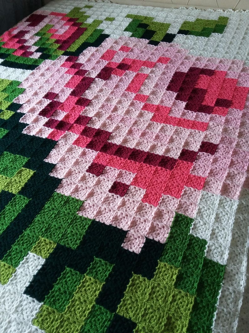 May include: A crocheted blanket with a pixelated design of a pink flower with green leaves. The blanket is made with different shades of pink, green, and white yarn.